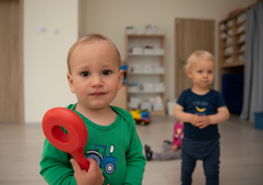Dwoje małych dzieci stoi w sali, jedno trzyma czerwoną zabawkę