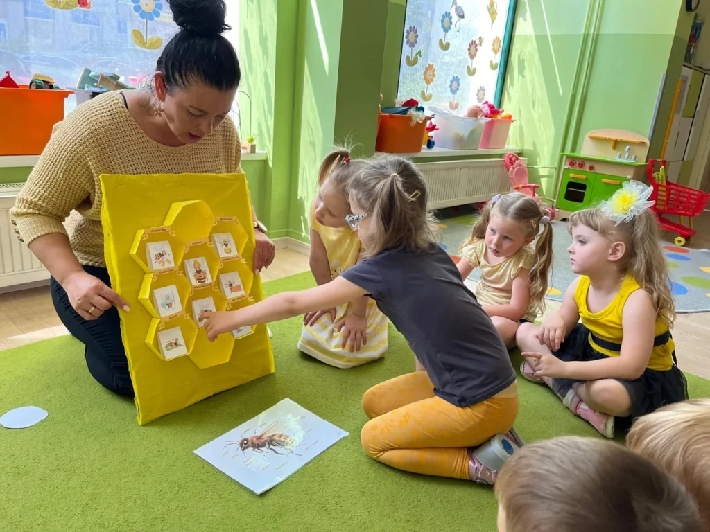 Nauczycielka pokazuje dzieciom żółtą tablicę w kształcie plastra miodu na zajęciach o pszczołach
