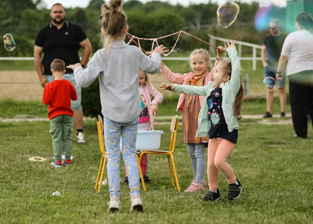 Dzieci bawią się na trawie bańkami mydlanymi, w tle dorośli