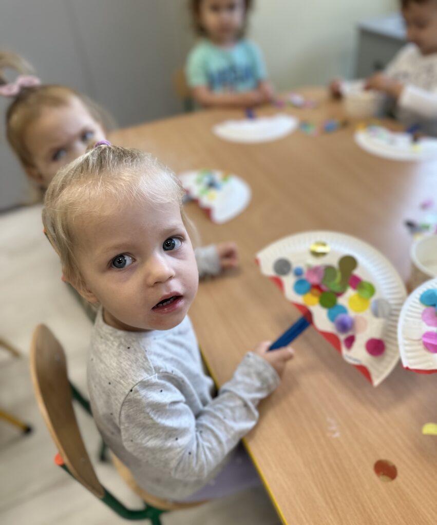 Dziewczynka przy stole robi prace plastyczne z talerzyków papierowych i kolorowych kółek