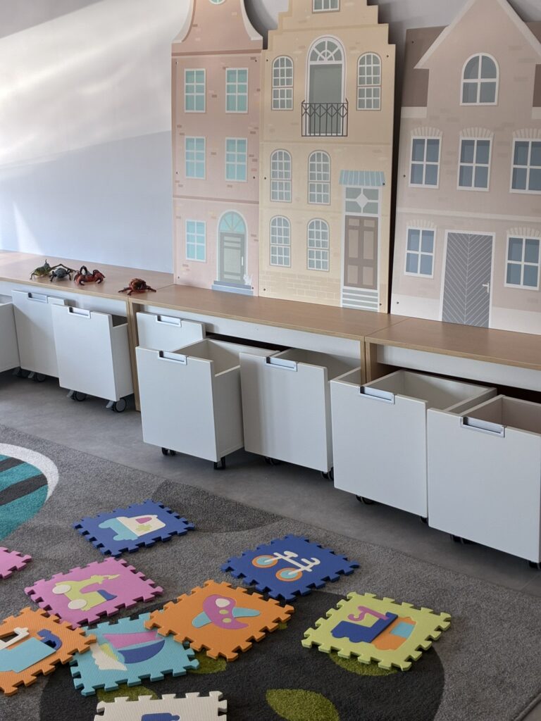 Kącik zabaw z piankowymi puzzlami na dywanie, białymi pojemnikami na kółkach i malowanymi domkami