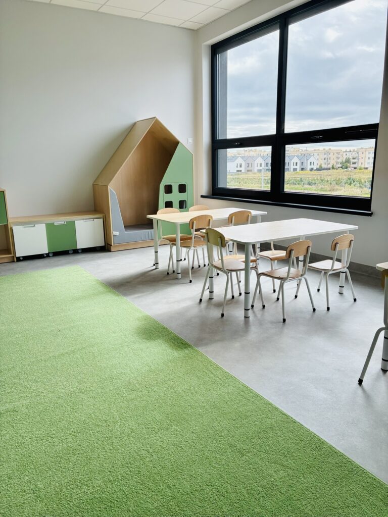 Sala z domkiem do czytania, stolikami z krzesełkami i zielonym dywanem przy dużym oknie
