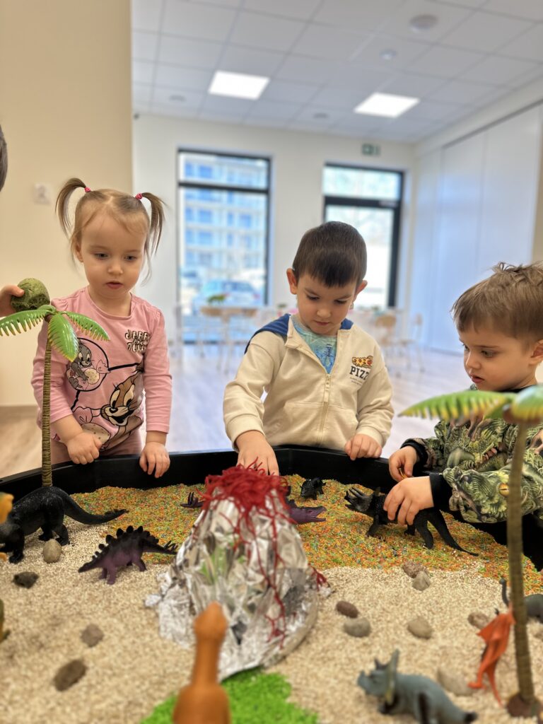Troje dzieci bawi się w sensorycznej tacy z dinozaurami, palmami i wulkanem
