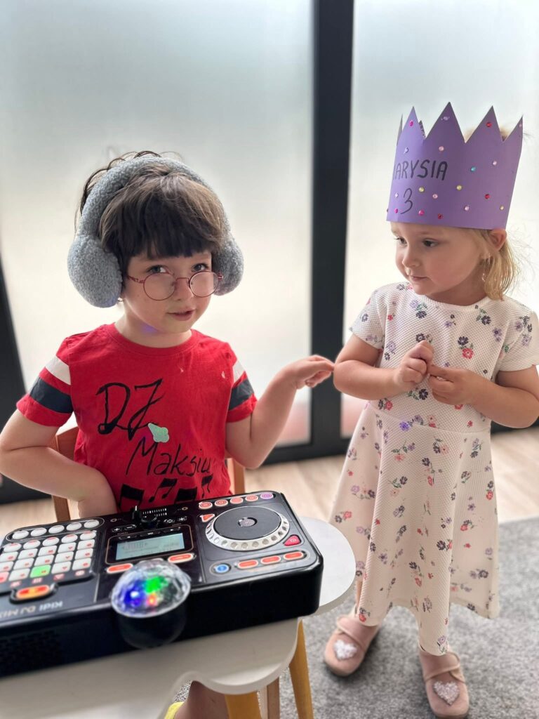 Dziecko w nausznikach i okularach przy zabawkowym mikserze DJ, obok dziewczynka w papierowej koronie