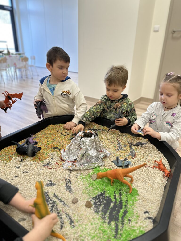 Dzieci bawią się przy tacy z piaskiem, wulkanem z folii i figurkami dinozaurów
