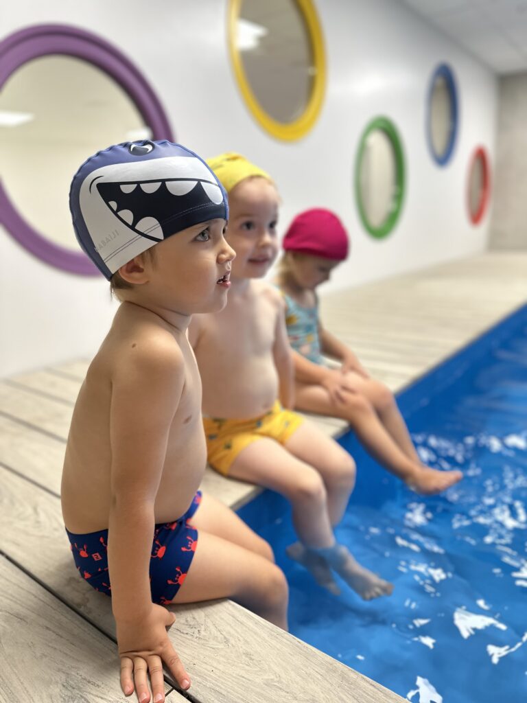 Troje dzieci w czepkach siedzi na brzegu basenu z nogami w wodzie