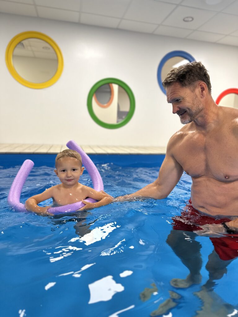 Dziecko z makaronem do pływania w basenie, obok stoi instruktor