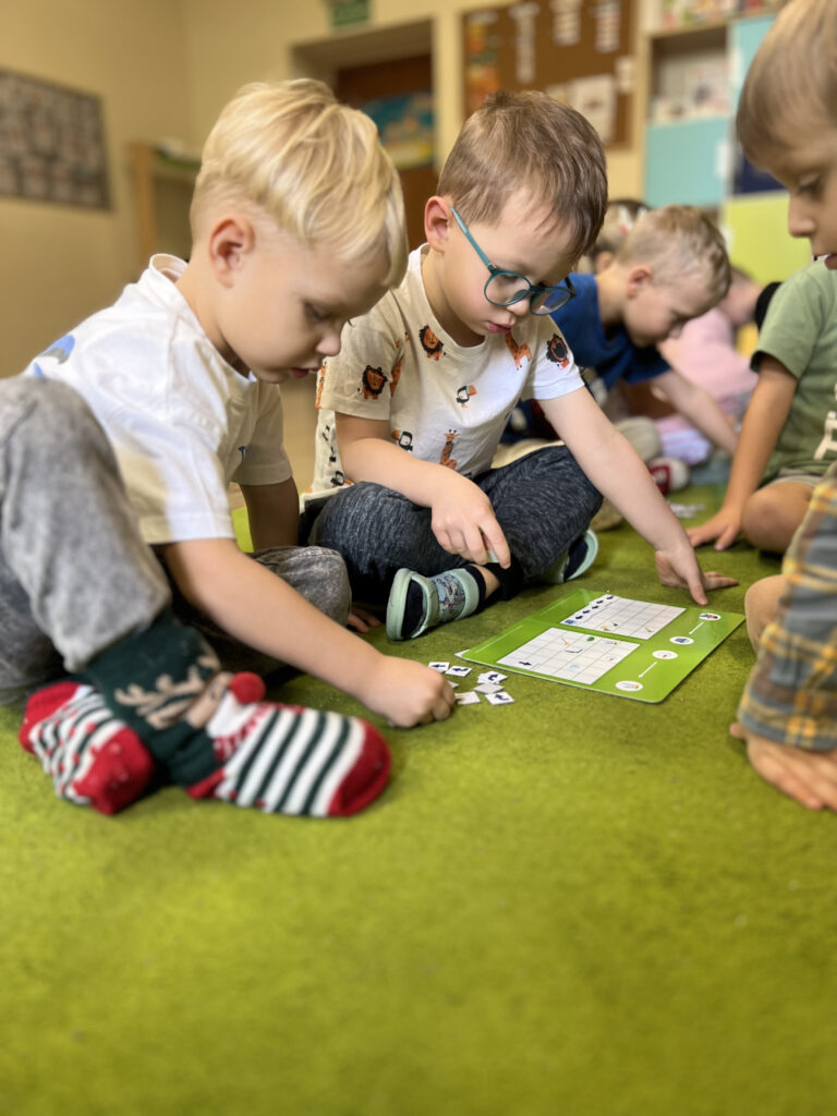 Dzieci siedzą na zielonym dywanie i rozwiązują zadanie z kartami